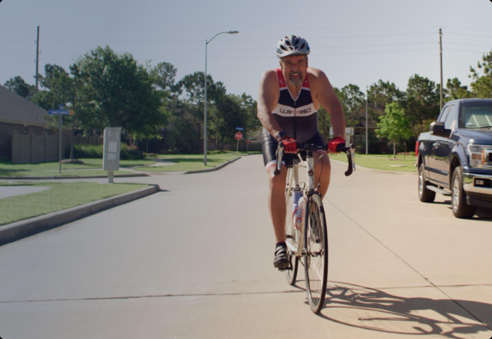 Cyclist in a helmet and racing attire rides on a suburban street, with trees and parked cars in the background on a sunny day.