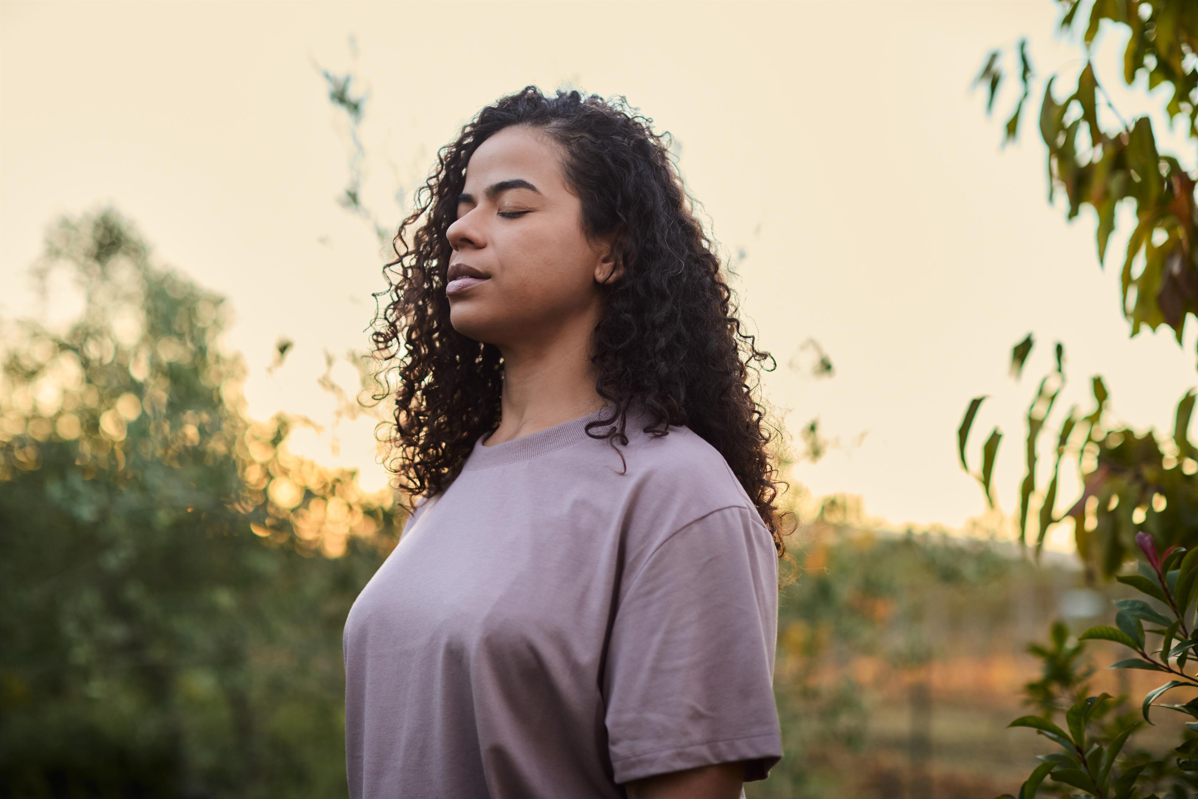Woman with curly hair stands outdoors, eyes closed, in a peaceful garden setting during sunset.