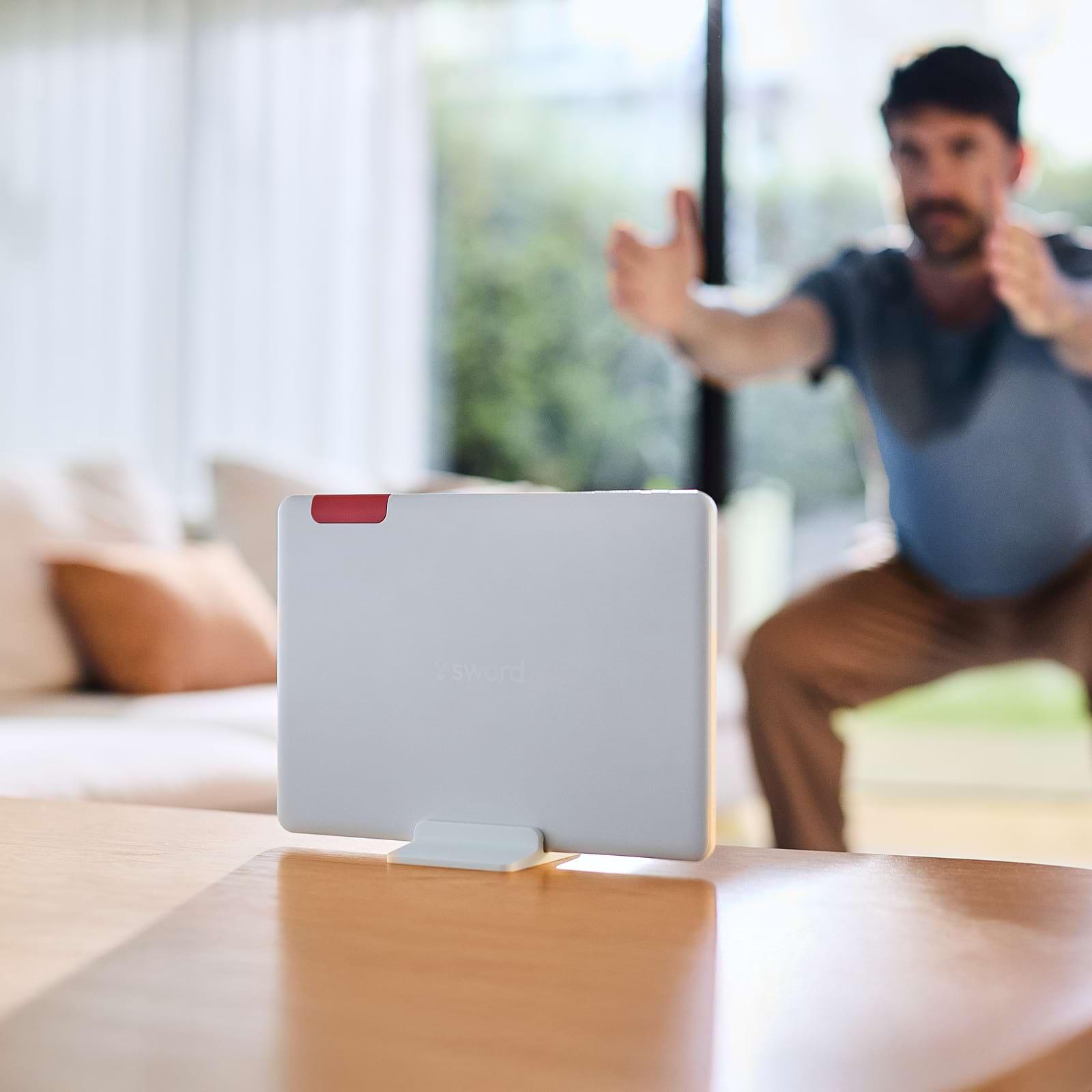 A person exercises indoors, facing a tablet on a stand. The room has a sofa and large windows with a view of greenery outside.