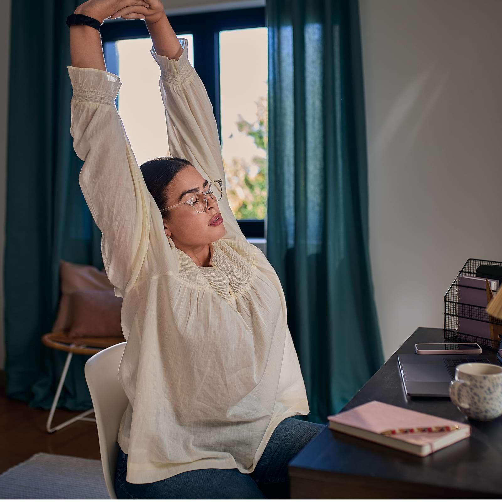 Person stretching at a desk with eyes closed, wearing glasses and a white blouse, surrounded by a laptop, notebook, and a mug.