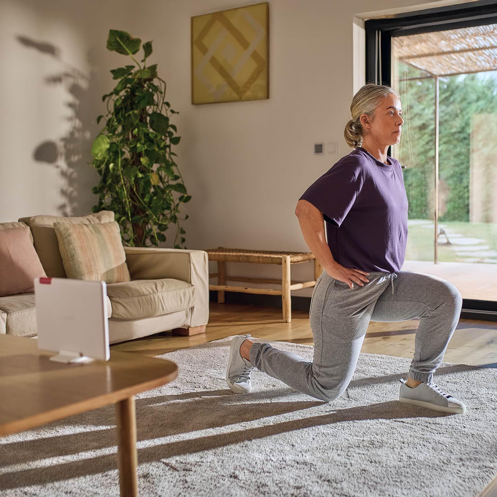 A woman in a purple shirt and gray pants performs a lunge in a cozy living room with a sofa, plant, and large window.