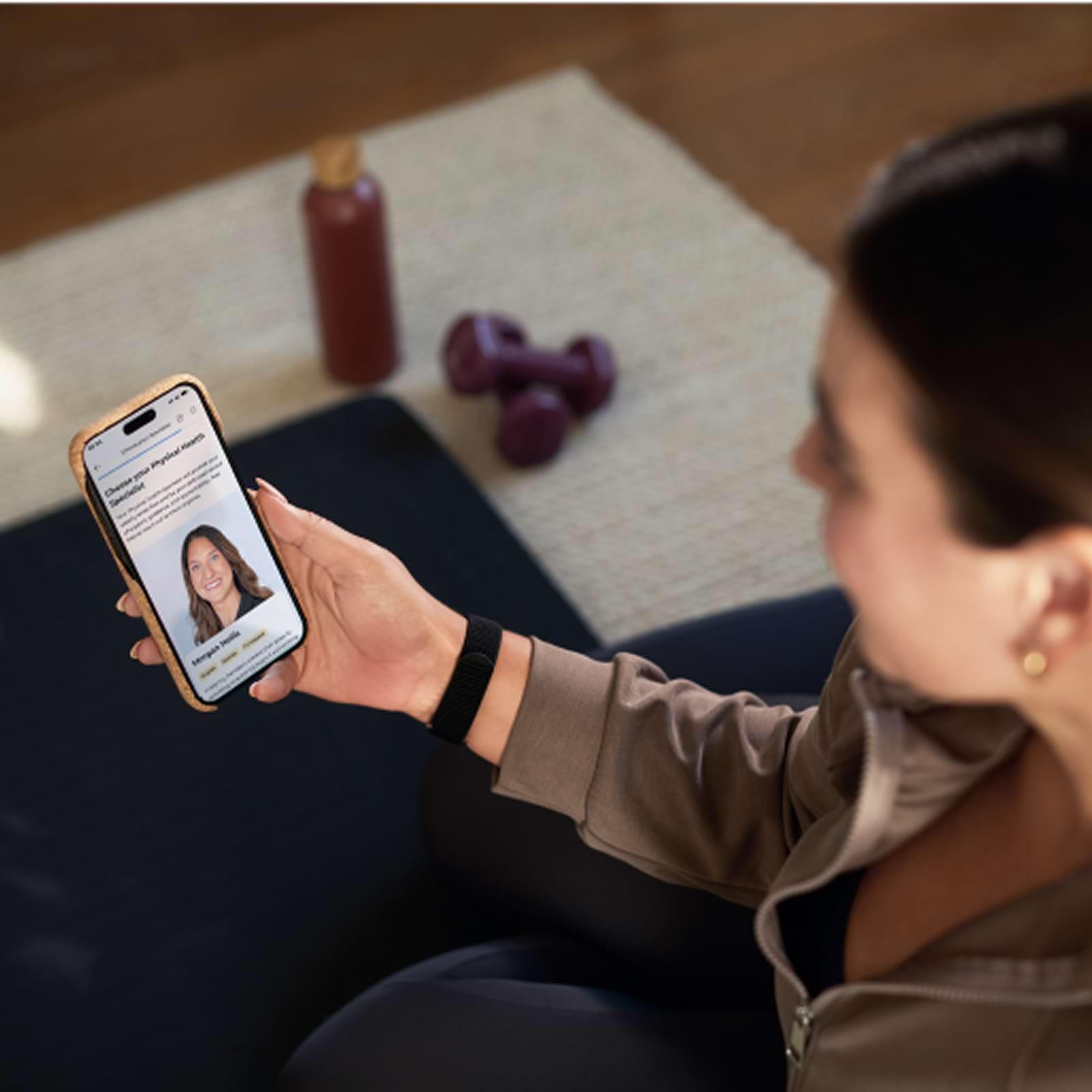 Person holding a smartphone displaying a video call, sitting on a mat with a water bottle and dumbbells nearby.
