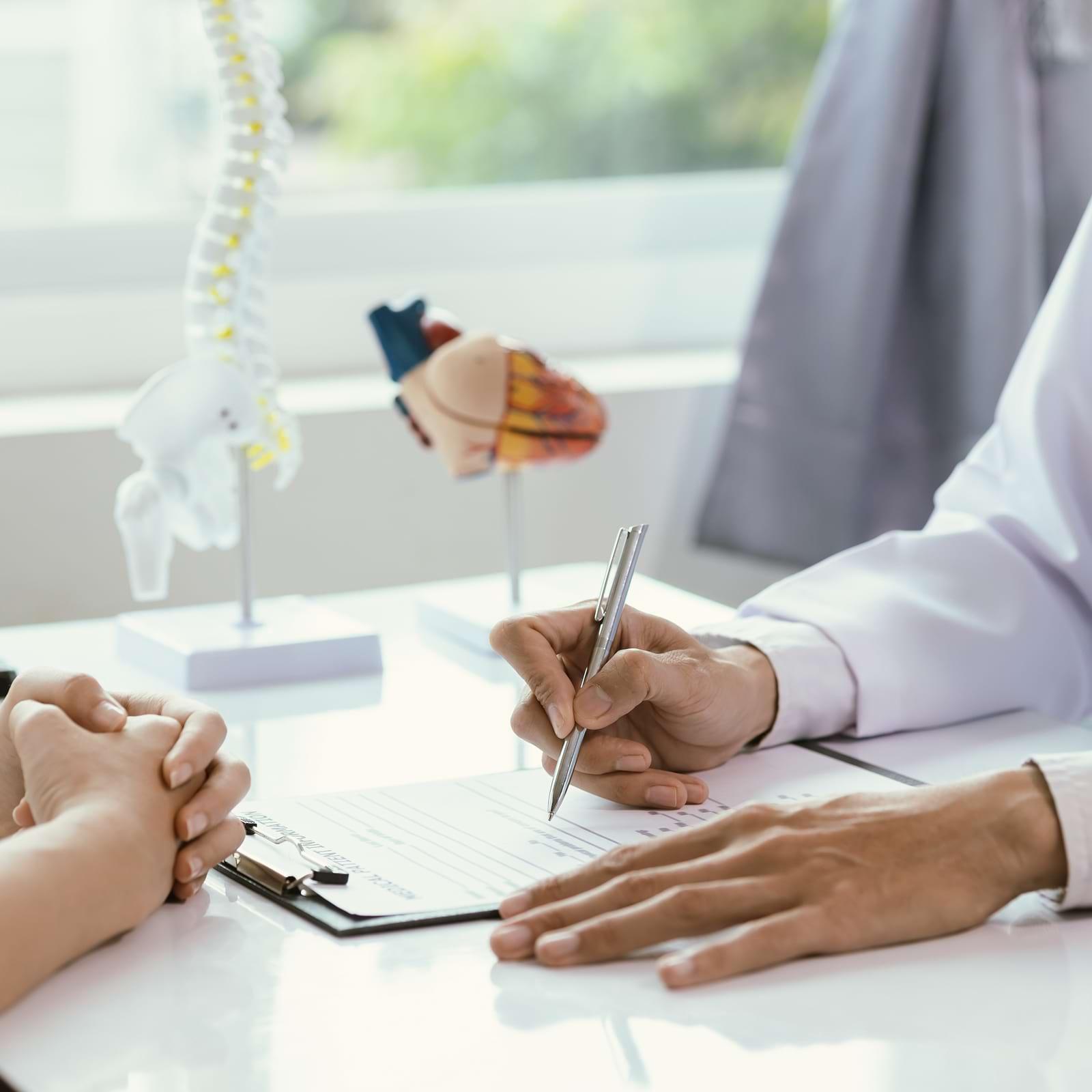 Doctor writing on a clipboard during a consultation, with anatomical models of a spine and heart in the background.