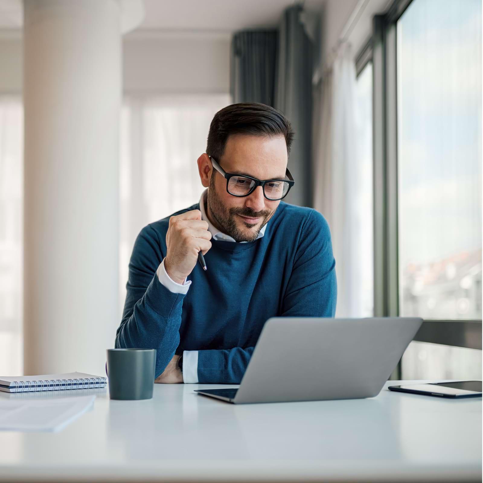 Man in glasses working on a laptop at a bright office desk, with a notebook, smartphone, and mug nearby.