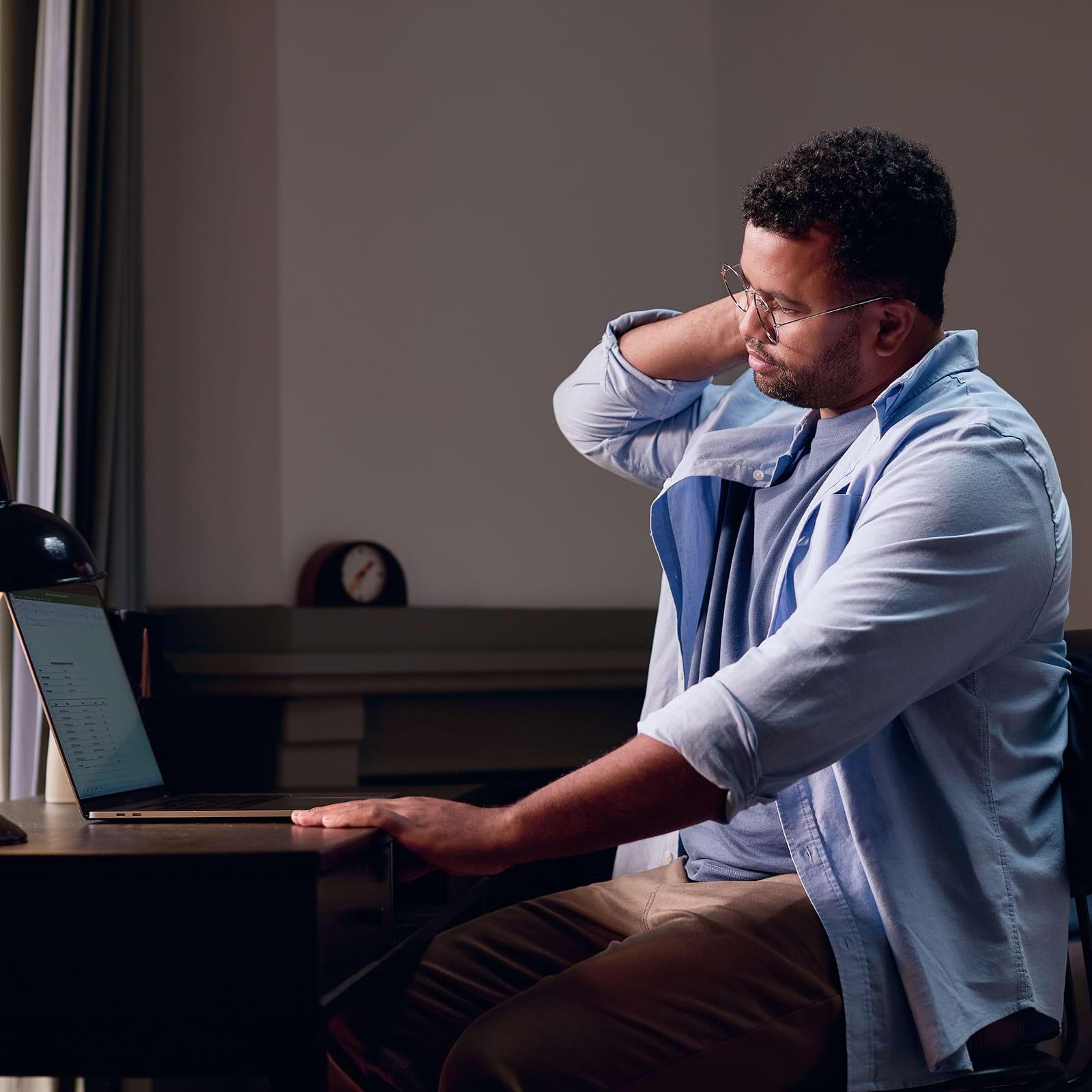 A man sitting at a desk, looking at a laptop, rubs his neck with one hand, appearing tired or tense.
