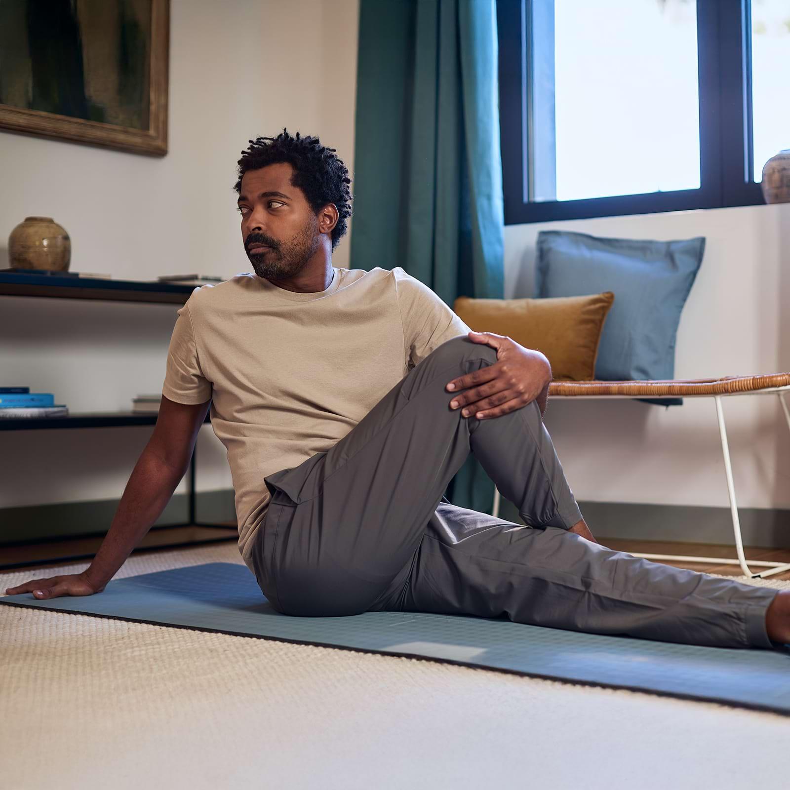 Man in a beige shirt and gray pants doing a seated twist yoga pose on a mat in a cozy room with pillows and a window.