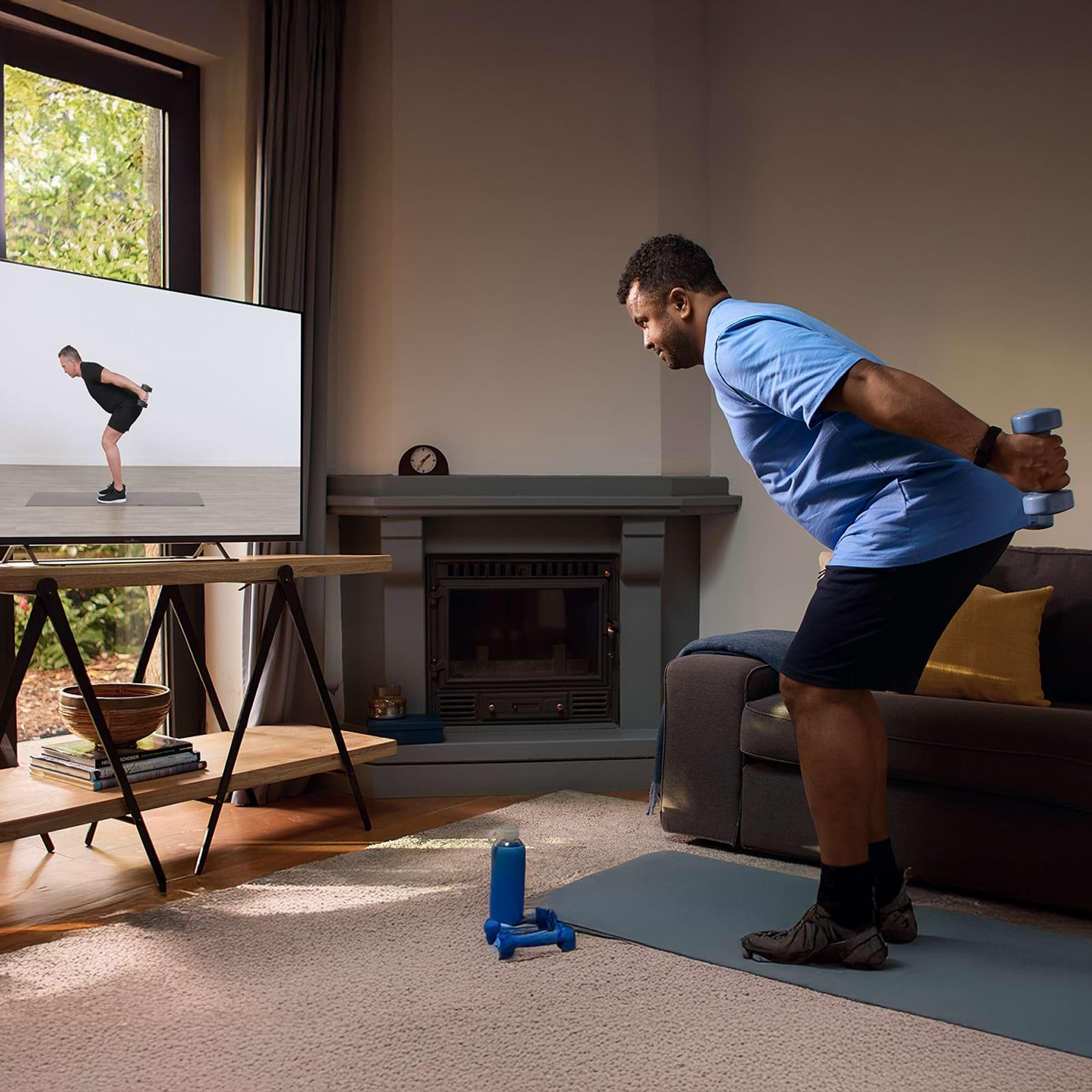 Man exercising at home with dumbbells, following a workout video on TV. He's on a mat in a cozy living room with a fireplace.