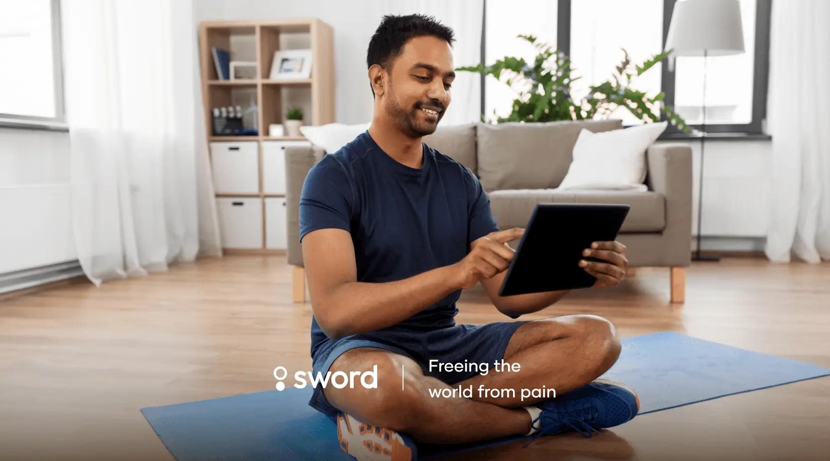 Man sitting cross-legged on a yoga mat in a living room, using a tablet. Text reads "Freeing the world from pain" with a logo.