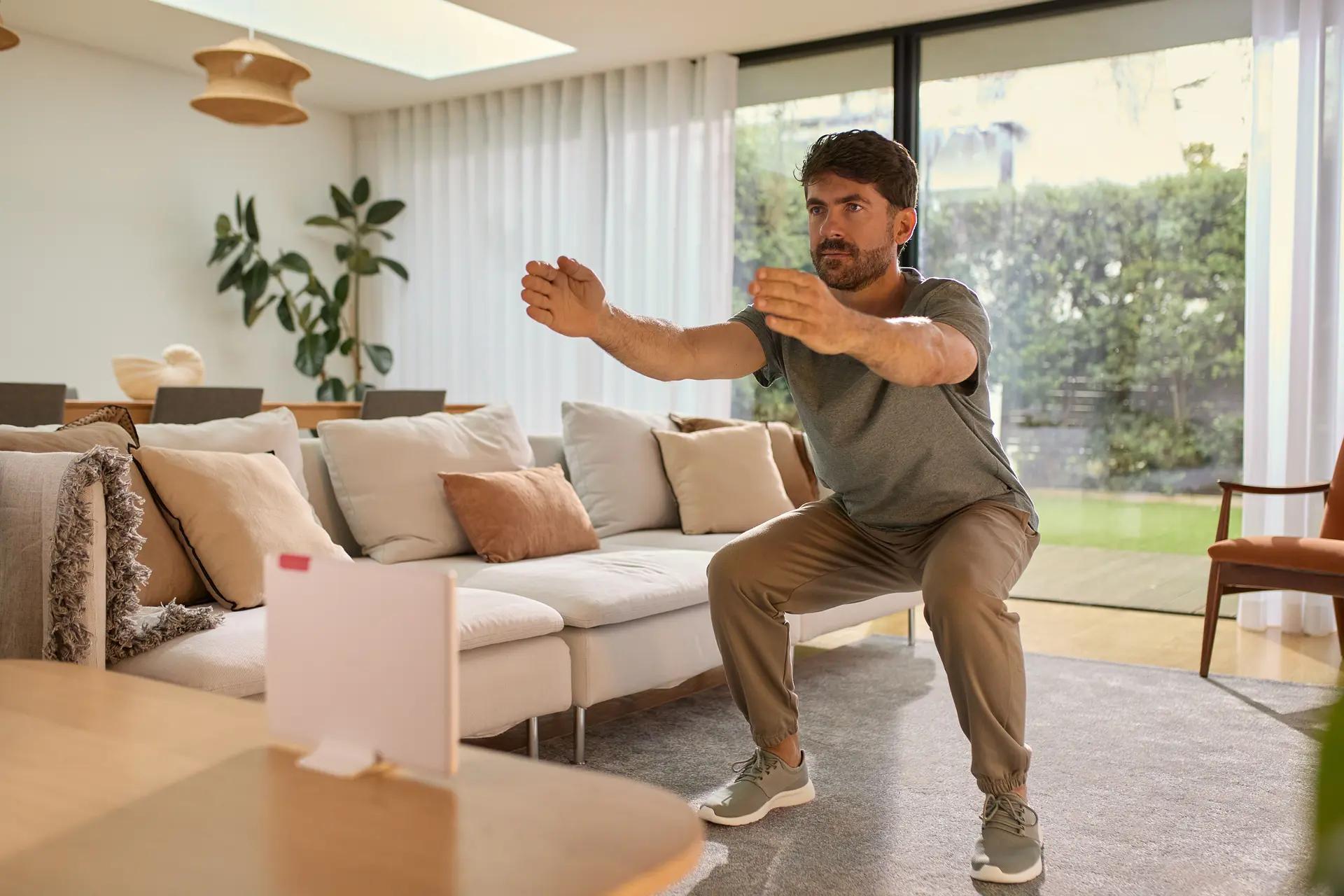 Man doing squats in a living room, wearing casual clothes. The room has a sofa, plants, and large windows with a view of greenery.