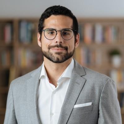 Profile image of Fernando Correia with glasses and a beard, wearing a gray suit and white shirt, standing in front of a blurred bookshelf background.
