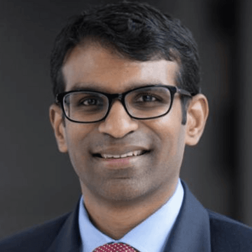 A person wearing glasses and a suit with a red tie, smiling against a blurred dark background.