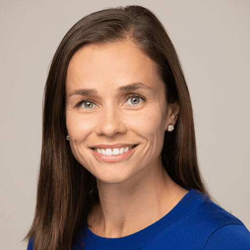 Smiling woman with long brown hair, wearing a blue top and silver earrings, against a neutral background.