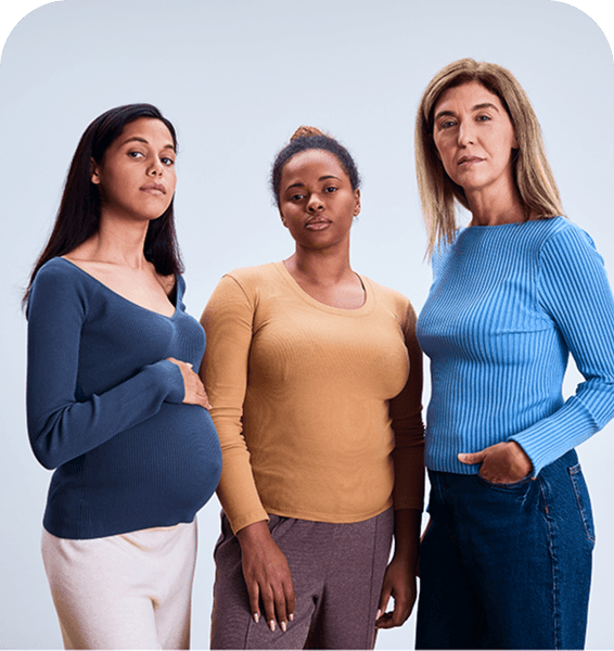 Three women standing together, each wearing different colored tops, with one woman visibly pregnant. Neutral background.